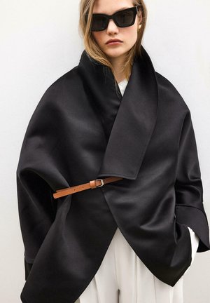 Vrouw draagt een oversized zwarte satijnen wikkeljas met een bruine riem, witte broek en zwarte zonnebril, staand tegen een effen witte achtergrond.