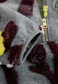 Gray fleece sweatshirt featuring textured floral patterns in burgundy, yellow, and green, with a gold zipper and gray cuffs.