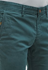 Teal bomullschinos med en mjuk textur, dragkedja, framfickor och en brun knapp med en orange accent nära fickstygn.