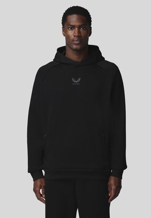Homme face à l'avant portant un sweat à capuche noir et un pantalon noir avec un logo blanc sur la poitrine et un fond gris uni.