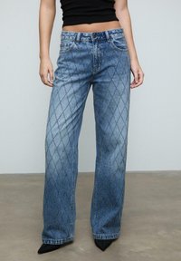 Ljusblå denimbyxor med diamantmönster och prydnader, rymlig passform och standard femfickdesign, tillsammans med spetsiga svarta skor.