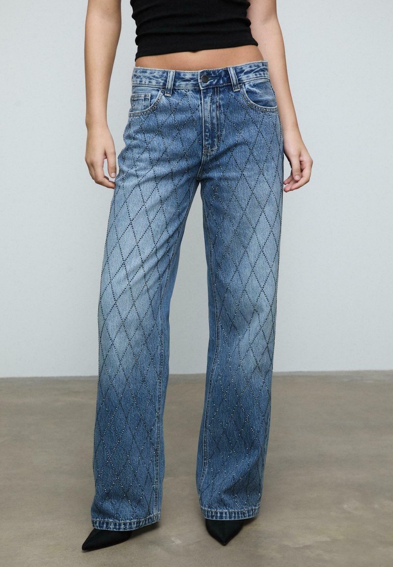 Ljusblå denimbyxor med diamantmönster och prydnader, rymlig passform och standard femfickdesign, tillsammans med spetsiga svarta skor.