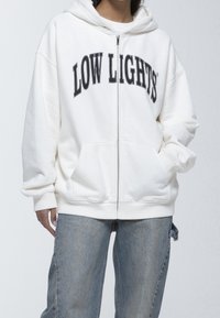 Sudadera blanca con cremallera y texto negro "LOW LIGHTS", material suave, amplios bolsillos frontales y un ajuste relajado con capucha ajustable.