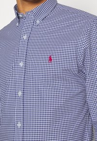 Košile s károvaným vzorem v námořnické modři a bílé, vyrobená z tkané látky. Má límec s knoflíky a malé červené logo na prsou.