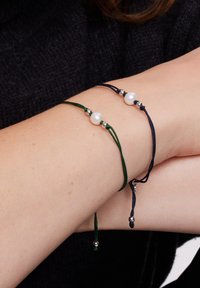 Dwa regulowane bransoletki, jedna zielona i jedna granatowa, z białymi perłami i srebrnymi koralikami, noszone na nadgarstku.