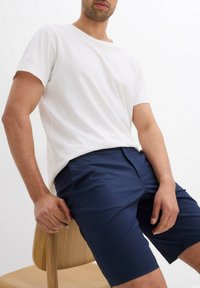 T-shirt bianca in cotone con scollo rotondo abbinata a pantaloni corti blu navy. Il design è semplice, con una vestibilità rilassata e una texture liscia, seduta su una sedia.