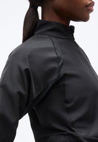 Κοντινό πλάνο ενός ατόμου που φοράει μαύρο μπουφάν με φερμουάρ και διχτυωτά πάνελ κάτω από τη μασχάλη, με χτένισμα κοτσίδας.