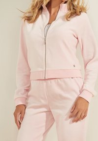 Sudadera de cremallera color rosa claro con cuello alto y puños acanalados, hecha de tela suave. Presenta un pequeño logo en la parte delantera y pantalones a juego.