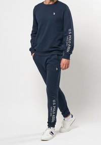 Mörkblå sweatshirt och byxor i set av bomull. Utrustad med vitt logotyp och text på ärmar och ben. Enkel, sportig design.