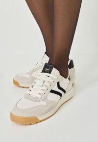 Beige Wildleder- und Ledersneaker mit schwarzen Akzenten, weißen Schnürsenkeln und einer strukturierten Gummisohle. Das Design weist gebogene Streifen an den Seiten auf.
