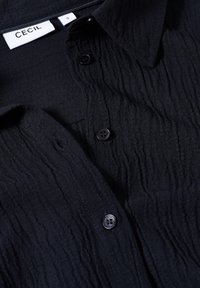 Chemise en tissu noir froissé avec des boutons et un col, affichant une étiquette de taille blanche marquée "S" et une étiquette de marque "CECIL".