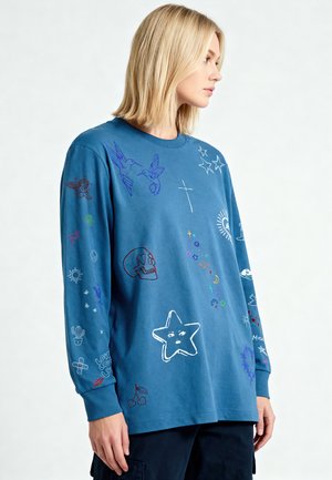 Personne blonde portant une chemise à manches longues bleue décorée de griffonnages colorés comprenant des oiseaux, des étoiles, un crâne, des cerises, un cactus et des motifs de soleil.