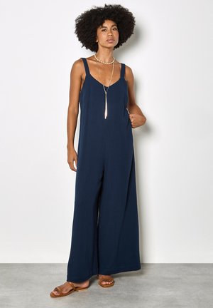 Kobieta o kręconych włosach, ubrana w granatowy, szerokie spodnie na ramiączkach oraz warstwowe złote naszyjniki, stojąca z jedną ręką w kieszeni.