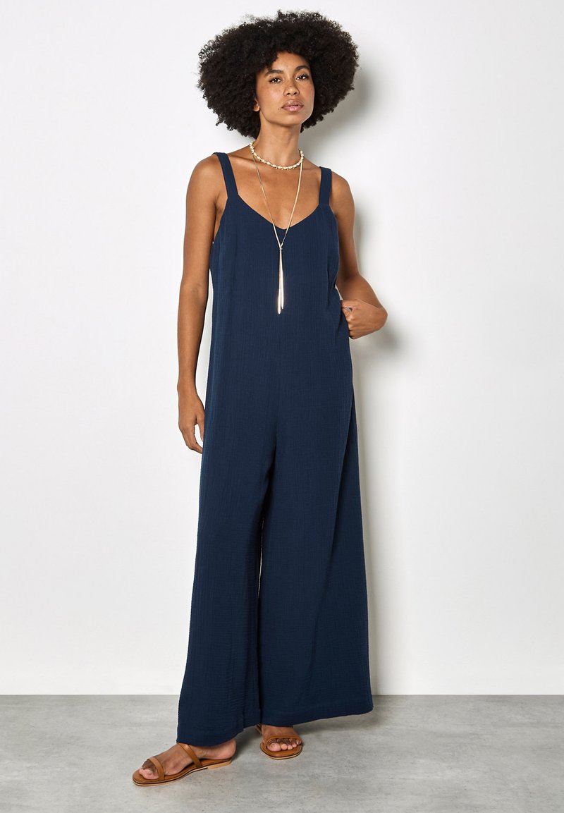 Kobieta o kręconych włosach, ubrana w granatowy, szerokie spodnie na ramiączkach oraz warstwowe złote naszyjniki, stojąca z jedną ręką w kieszeni.