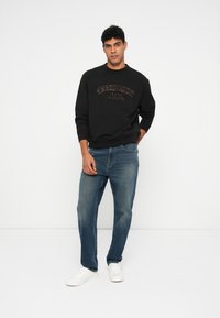 Čierna mikina s logom "Tommy Jeans" v hnedých písmenách, skombinovaná s modrými džínsami a bielymi teniskami. Pohodlný outfit s jednoduchým dizajnom.