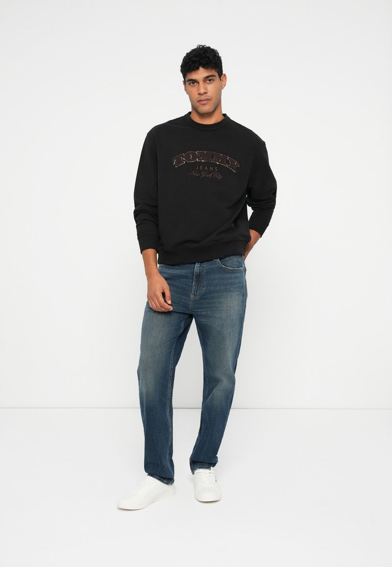 Čierna mikina s logom "Tommy Jeans" v hnedých písmenách, skombinovaná s modrými džínsami a bielymi teniskami. Pohodlný outfit s jednoduchým dizajnom.