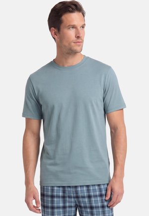 Mann trägt ein hellblaues kurzärmeliges T-Shirt und blaue karierte Hose, steht und schaut nach rechts.