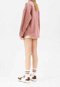 Różowa, oversize'owa kurtka dżinsowa z długimi rękawami, bocznymi rozcięciami i zapięciem na guzik. Nosi się ją na białe szorty z postrzępionymi krawędziami oraz brązowymi trampkami.