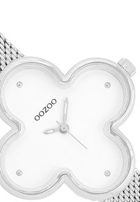 Montre avec bracelet en maille argentée et un cadran blanc en forme de trèfle, comportant des index horaires et des aiguilles noires. Nom de la marque "OOZOO" sur le cadran.