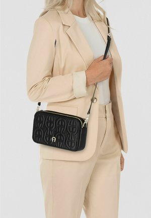 Zwarte schoudertas met doorgestikt patroon en gouden hardware, voorzien van een ritssluiting en verstelbare band. Draagt een beige pak.