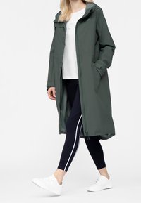 Lang, olivengrøn let regnfrakke med hætte, sidelommer og lynlås, båret over en hvid skjorte og marineblå leggings med hvide striber.