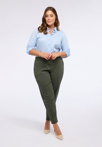 Camicia azzurro chiaro con maniche arrotolate, abbinata a pantaloni slim-fit verde oliva con tasche laterali e tacchi beige.