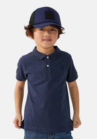 Donkerblauwe polo met drie knopen, gestructureerde stof en een klein logo. Gedragen met een blauwe pet met een zwart patch, gecombineerd met denim shorts.