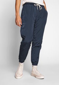 Person klädd i marinblå nylonjoggingbyxor med elastiska muddar och vita högstövlar, stående mot en enfärgad bakgrund.