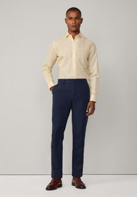 Camicia giallo chiaro con bottoni e tasca, abbinata a pantaloni blu navy e scarpe in pelle marroni. La texture del tessuto appare liscia e fresca.