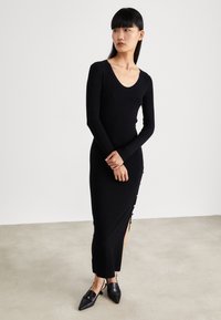 Black ribbed knit dress with long sleeves, deep V-neck, and side slit featuring button accents. Slim fit, floor-length design.