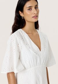 White dress made of textured fabric with embroidered patterns and cut-out details. Features a v-neck and short flared sleeves.