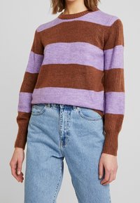 Randig tröja i brunt och lavendel, tillverkad av mjukt stickat material. Har en rundad halsringning och ribbade ärmslut, kombinerat med ljusblå denimjeans.