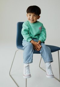 Minzgrüner Pullover mit pinkem Palmenmotiv, kombiniert mit blau-weiß gestreiften Hosen und weißen Turnschuhen. Sitzend auf einem blauen Stuhl.