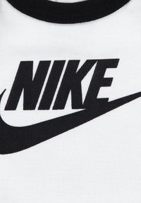 Tissu blanc avec un col en côtelé noir et un grand logo "NIKE" noir avec un swoosh. Le matériau semble lisse avec un motif texturé.