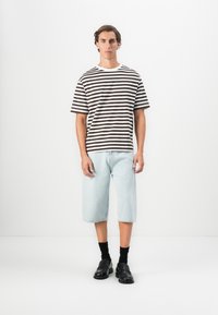 Mann iført svart og hvitt stripet t-skjorte, lyseblå denimshorts til kneet, svarte sokker og svarte loafers som står foran en hvit bakgrunn.