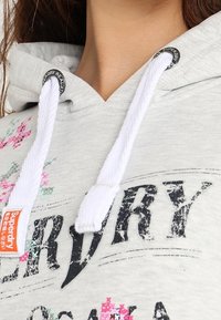 Sudadera de algodón gris con cordones blancos, que presenta un gráfico prominente en negro y acentos florales de colores. Textura suave, diseño casual.