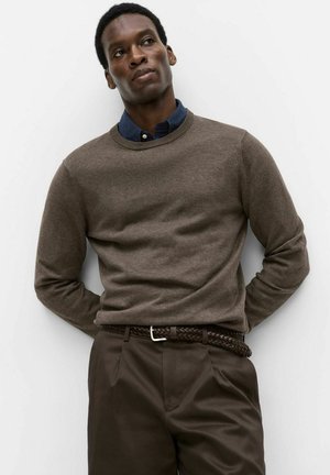 Mand iført en brun sweater over en blå skjorte med krave, mørkebrun flettet bælte og mørkebrune bukser, stående mod en hvid væg.