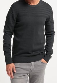 Schwarzer, strukturierter Strickpullover mit gerippten Bündchen und Saum. Verfügt über ein horizontales Streifenmuster auf der Brust. Kombiniert mit grauen Jeans.