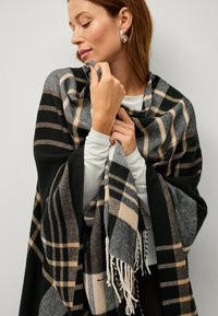Wool plaid blanket scarf in black, beige, and gray tones with fringed edges, draped over shoulders, showcasing a soft texture.