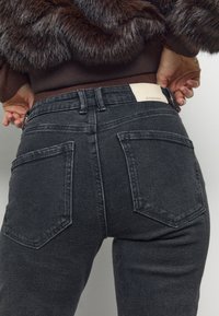 Musta korkeavyötäröinen farkut, joissa on sileä pinta, kaksi takataskua, nahkainen merkintä vyötäröllä ja suora siluetti.