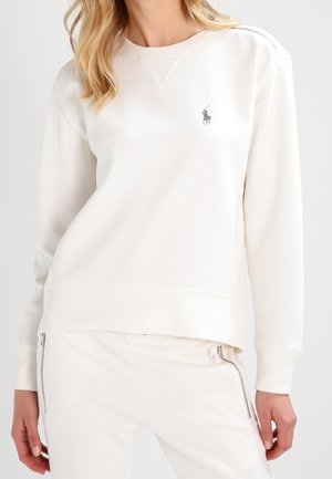 Nainen pukeutunut valkoiseen pitkähihaiseen svetariin ja vastaaviin valkoisiin housuihin, joissa on etutaskujen vetoketjut, seisoo rento asento käsivarret rentoina.