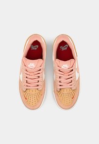 Ténis Nike em rosa claro e camurça bege; apresenta detalhes em branco, atacadores rosa, caixa de ponta perfurada e detalhes do logótipo em vermelho.