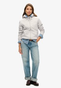 Sudadera gris claro con cremallera y capucha ajustable, llevada sobre una camisa de denim, combinada con unos vaqueros holgados de color azul claro y zapatos burdeos.