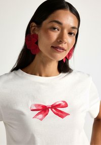 Biała bawełniana koszulka z nadrukiem czerwonej kokardy w akwareli na środku. Modelka ma na sobie duże czerwone kwieciste kolczyki oraz proste, ciemne włosy.