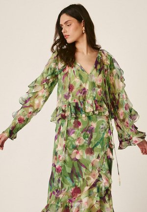 Mujer con un vestido largo de manga larga, verde con flores transparentes y volantes, mirando hacia su izquierda con los brazos ligeramente extendidos.