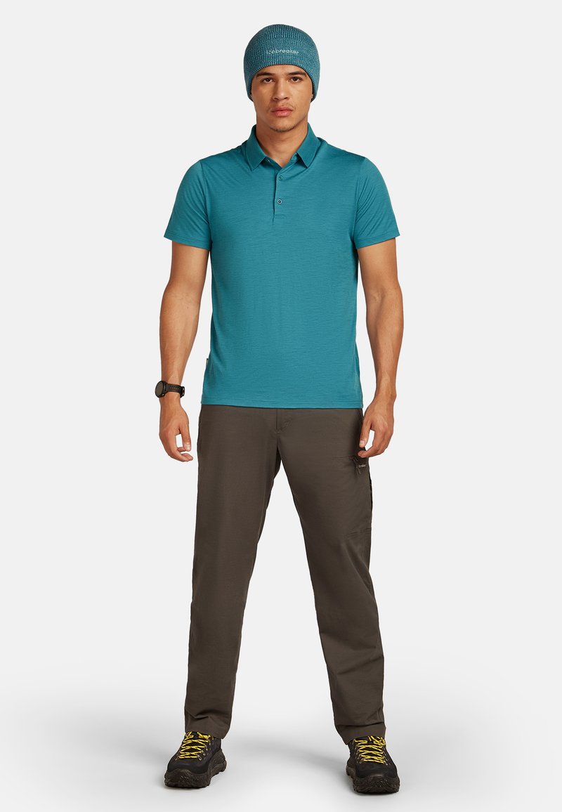 Turkos polo skjorta, lättvikts, kort ärm, tvåknapps placket. Mörkgrå cargobyxor med flera fickor. Svarta vandringskängor. Stickad mössa.