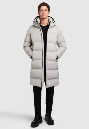 Lange, lichtgrijze puffercoat met een capuchon. Gequilt ontwerp, zijzakken en een label op de onderste zoom. Gepaard met zwarte broeken en laarzen.