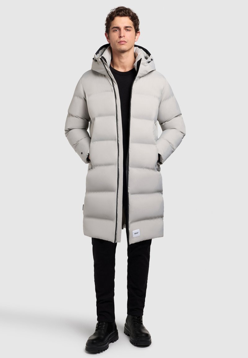 Lang lysegrå pufferjakke med hætte. Quiltet design, sidelommer og et mærke på den nedre kant. Sammen med sorte bukser og støvler.