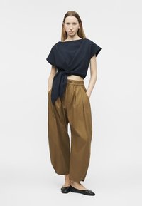 Frau steht mit den Händen in den Taschen, trägt ein schwarzes, geknotetes Crop-Top, lockere braune Hosen und schwarze Ballerinas.