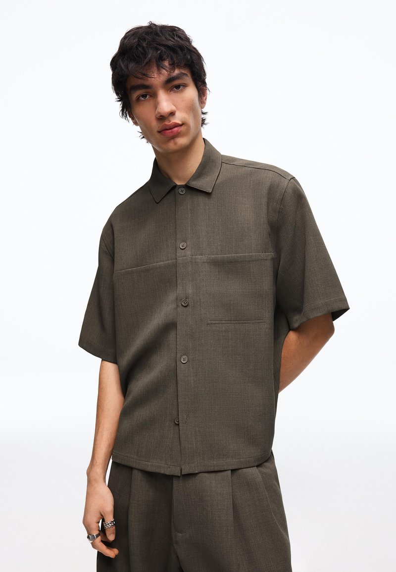 Ung mand med mørkt krøllet hår iført en løs olivengrøn kortærmet skjorte med knapper og matchende bukser mod en ensfarvet hvid baggrund.